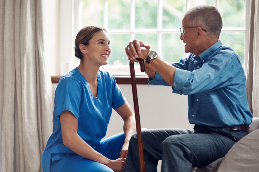 A smiling caregiver in blue scrubs sits with a senior man holding a wooden cane, sharing a warm conversation near a sunlit window.