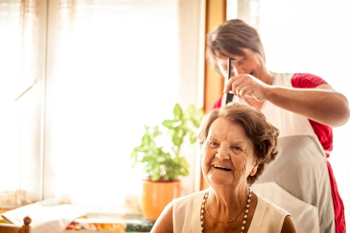 Caucasian senior woman has her hair styled. She is sitting in the foreground with the stylist to the back of her. She has short brown, wavy hair and is wearing a sleeveless, light cream linen top, a pearl necklace and short gold necklace. She is smiling. The stylist is behind her with a comb and is wearing a red top and apron. She has short brown hair. In the background is a large window and plant in a yellow pot.