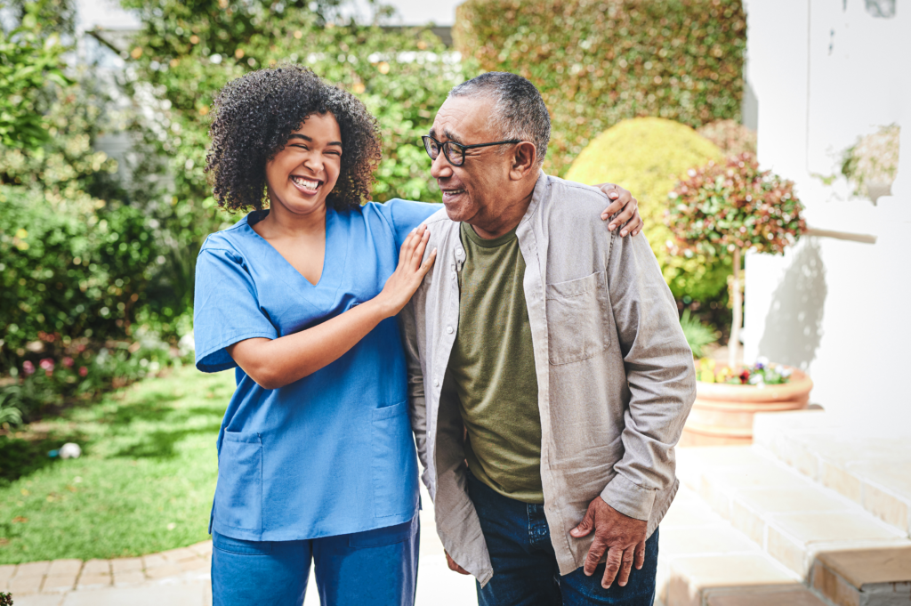 A smiling caregiver in blue scrubs walks alongside a senior man in a garden, with her hand supportively on his shoulder
