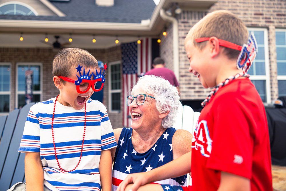 A smiling grandmother in a star-spangled top laughs with her two grandsons in patriotic fourth of july attire on a porch decorated with an american flag.